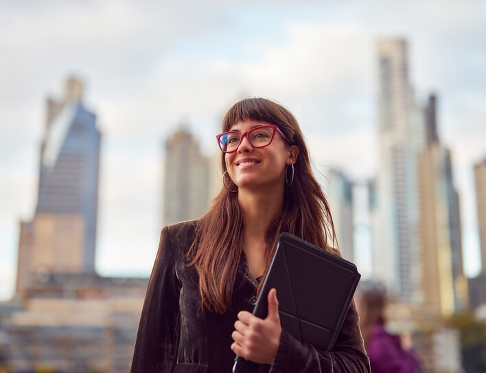 Eine junge Frau mit Brille und Tablet steht lächelnd vor einer modernen Skyline und blickt optimistisch in die Ferne.