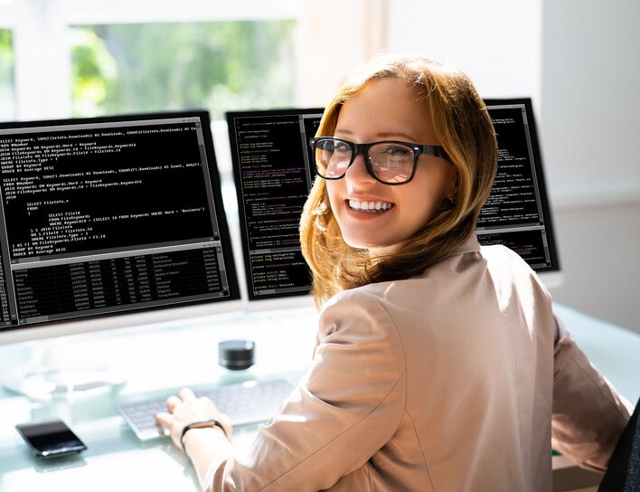 Lächelnde Frau sitzt vor zwei Bioldschirmen, auf denen Code zu sehen ist.