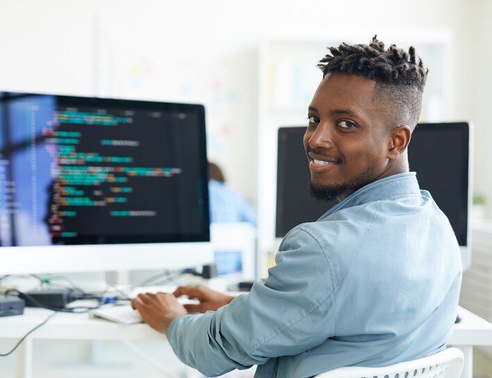 Freundlicher Mann sitzt am Schreibtisch vor einem Bildschirm mit Code und schaut in die Kamera