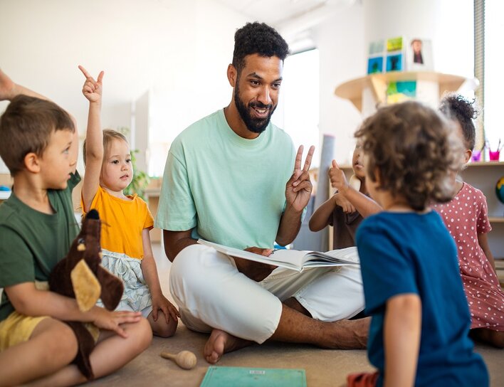 Ein junger Erzieher liest einer Gruppe von vier kleinen Kindern in einem hellen Raum aus einem Buch vor.