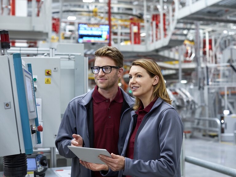 Eine Frau und ein Mann in Arbeitskleidung stehen in einer Produktionshalle, betrachten gemeinsam ein Tablet und überwachen dabei eine industrielle Anlage an einem Bedienpanel.