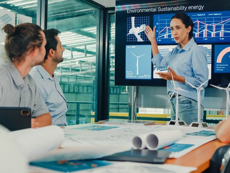 Eine Ingenieurin präsentiert einem Team nachhaltige Energiekonzepte mit Windkraftmodellen und Datenvisualisierungen.