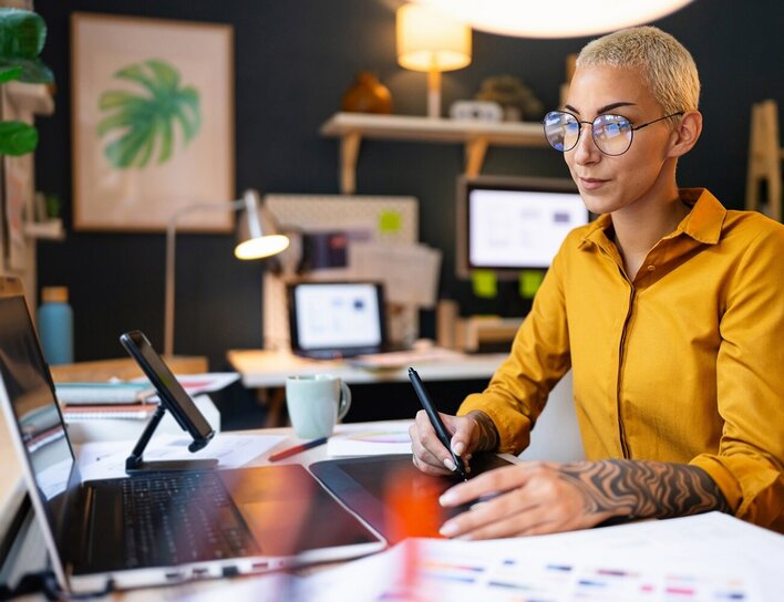 Frau mit Brille und kurzem blondem Haar arbeitet mit Grafiktablett und Laptop in einem modern eingerichteten Homeoffice.