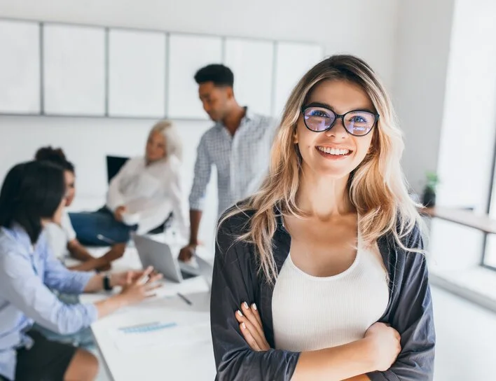 Frau mit Brille steht lächelnd vor einem Team, das in einem modernen Büro an einem Besprechungstisch arbeitet.