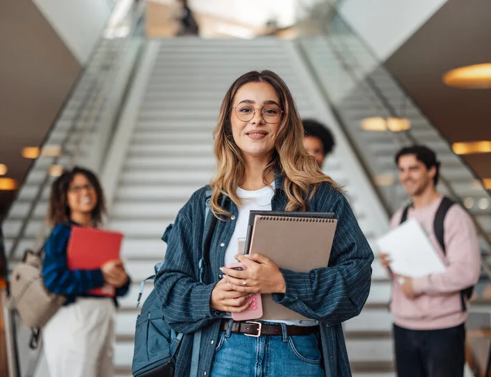 Junge lächelnde Studentin mit Brille und Notizbuch im Vordergrund vor einer Campus-Treppe mit weiteren Studierenden.