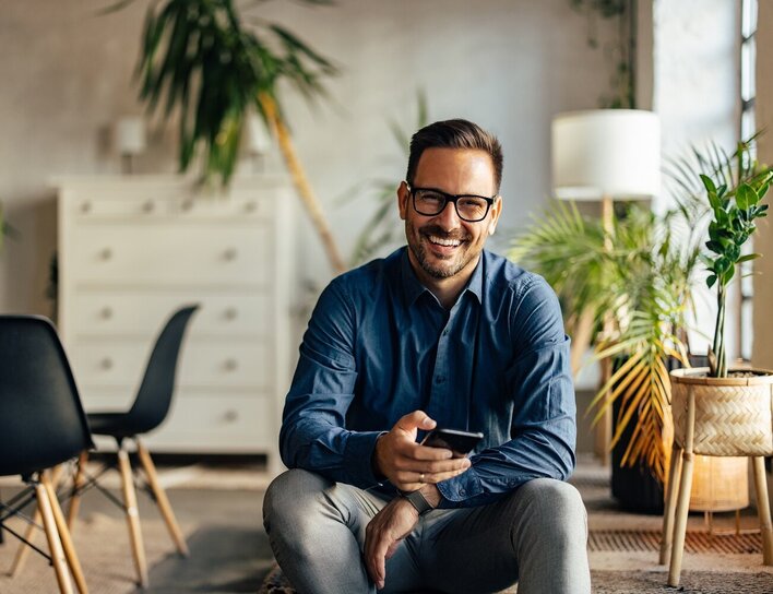 Lächelnder Mann mit Brille und Smartphone Lächelnder Mann mit Brille und Smartphone sitzt entspannt auf einem Sofa in einem modern eingerichteten Wohnzimmer mit Pflanzen und stilvollen Möbeln.