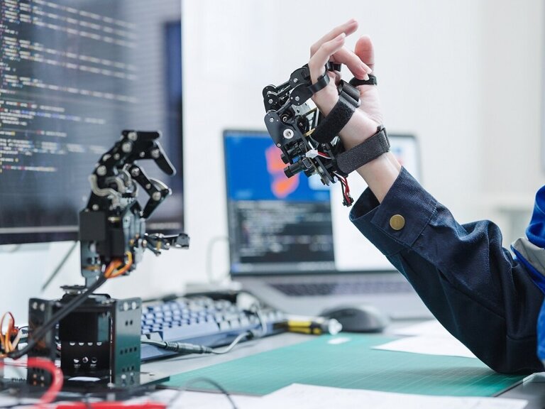 Eine Person testet ein robotisches Hand-Exoskelett vor einem Bildschirm mit Programmcodes in einem modernen Technikumfeld.