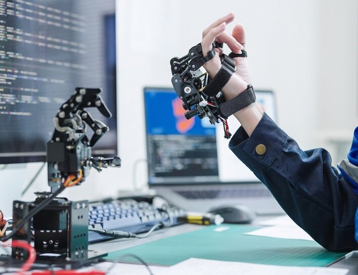 Eine Person testet ein robotisches Hand-Exoskelett vor einem Bildschirm mit Programmcodes in einem modernen Technikumfeld.