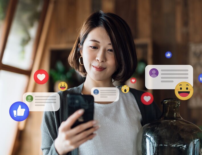 Eine junge Frau blickt lächelnd auf ihr Smartphone, während animierte Social-Media-Symbole wie Likes und Emojis das Bild umgeben.