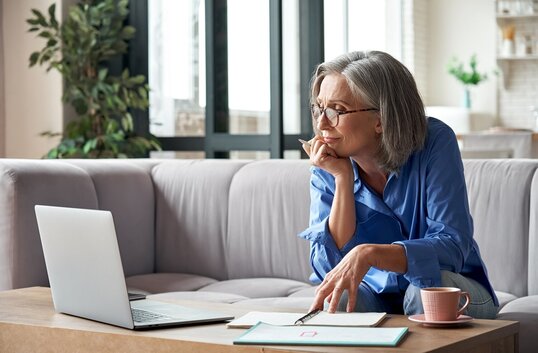 Frau sitzt auf dem Sofa und arbeitet mit Laptop, Notizbuch und Kaffeetasse im Wohnzimmer.