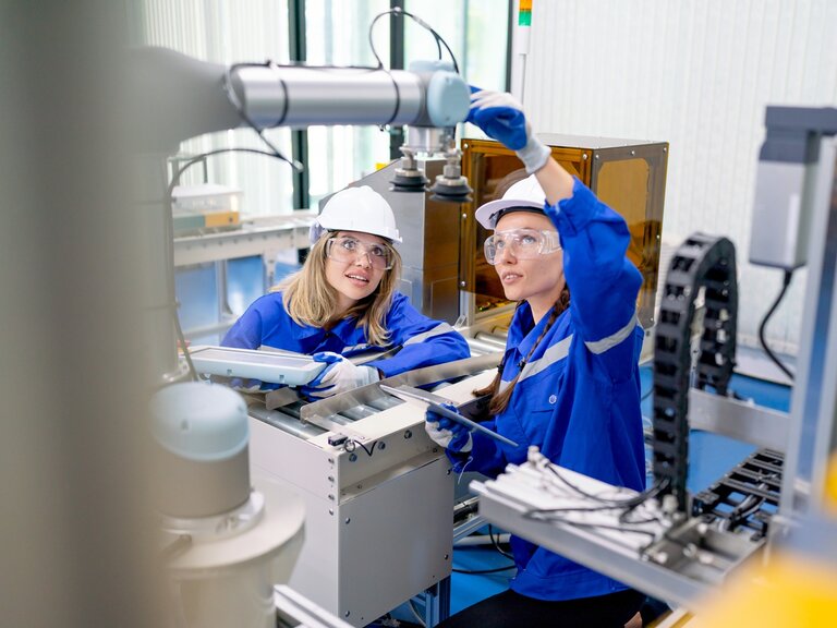 Eine Frau mit Schutzhelm und Schutzbrille justiert gemeinsam mit einer Kollegin einen Industrieroboter, während beide Tablets in einer modernen Produktionsumgebung nutzen.