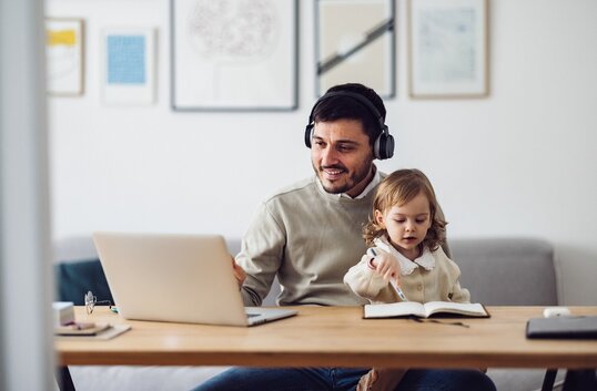 Vater im Homeoffice mit Laptop und Headset, neben ihm seine Tochter mit einem Buch.