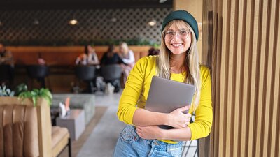 Eine junge Frau mit Brille und Mütze steht in einem gemütlichen Café, hält einen Laptop im Arm und lächelt freundlich in die Kamera.