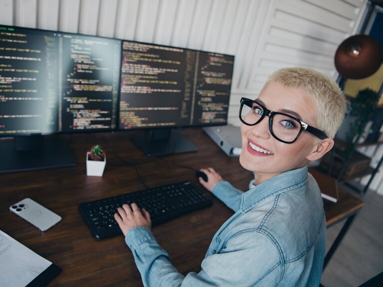 Eine lächelnde Frau sitzt vor mehreren Monitoren mit Programmiercode in einem modernen Arbeitsumfeld.