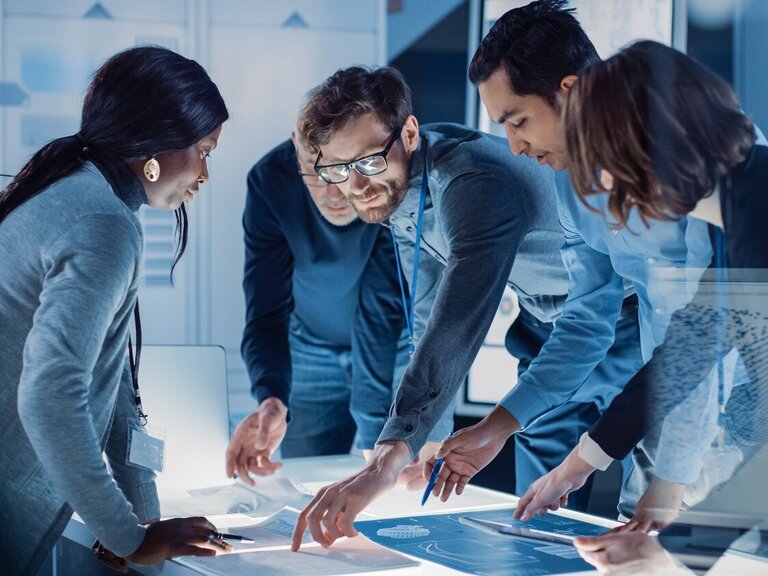 Ein interdisziplinäres Team diskutiert gemeinsam an einem digitalen Tisch über technische Entwürfe in einem modernen Innovationsumfeld.