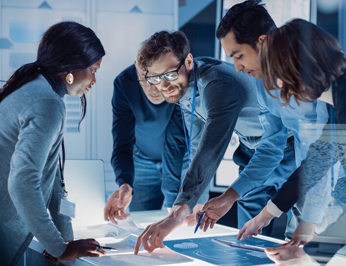 Ein interdisziplinäres Team diskutiert gemeinsam an einem digitalen Tisch über technische Entwürfe in einem modernen Innovationsumfeld.