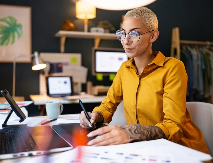 Frau mit Brille und kurzem blondem Haar arbeitet mit Grafiktablett und Laptop in einem modern eingerichteten Homeoffice.