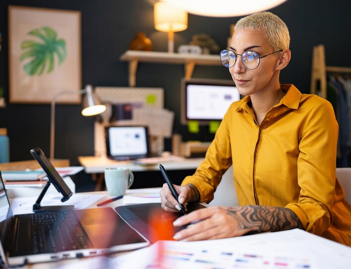 Frau mit Brille und kurzem blondem Haar arbeitet mit Grafiktablett und Laptop in einem modern eingerichteten Homeoffice.