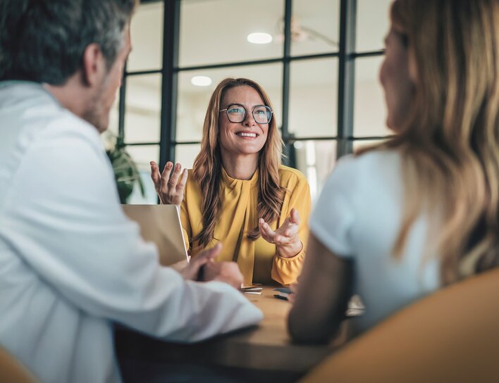 Eine Frau in gelber Bluse sitzt an einem Tisch und spricht engagiert mit zwei Personen, die ihr gegenübersitzen. Die Szene spielt in einer modernen, hellen Büroumgebung und vermittelt den Eindruck eines professionellen Beratungsgesprächs im Gesundheitssektor. Ein Laptop und Unterlagen liegen auf dem Tisch.
