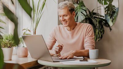 Ein junger Mann sitzt entspannt an einem Tisch mit Laptop, Unterlagen und Kaffeetasse, lächelt konzentriert in Richtung Bildschirm und ist von Zimmerpflanzen umgeben.
