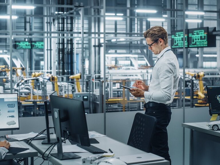 Zwei Personen arbeiten an technischen Visualisierungen in einem modernen Büro mit Blick auf eine automatisierte Produktionshalle.