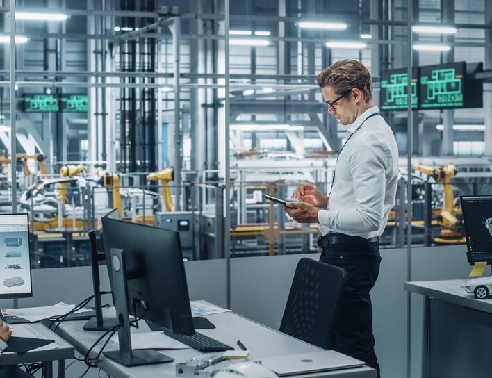 Zwei Personen arbeiten an technischen Visualisierungen in einem modernen Büro mit Blick auf eine automatisierte Produktionshalle.