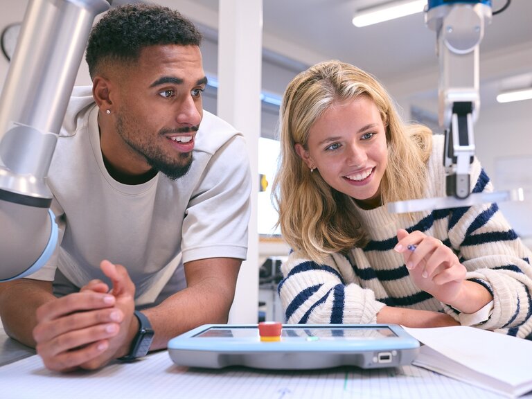Eine Frau in gestreiftem Pullover und ein Mann stehen an einem Labortisch und beobachten gemeinsam einen kleinen Industrieroboter, während sie über ein Steuergerät und Notizen sprechen.