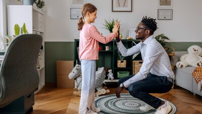 Ein Mädchen steht einem hockenden Mann gegenüber, beide lächeln sich an und geben sich ein High-Five. Sie befinden sich in einem freundlich eingerichteten Raum mit Spielzeug und kindgerechter Ausstattung.
