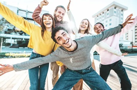 Gruppe junger Menschen, die draußen lachend in die Kamera posieren, mit ausgestreckten Armen und fröhlichen Gesichtern vor städtischer Kulisse.