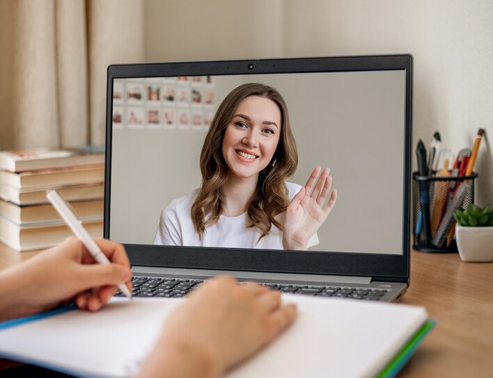 Virtuelle Kommunikation im Videochat Das Bild zeigt eine junge Frau, die auf dem Bildschirm eines Laptops freundlich in die Kamera lächelt und winkt. Im Vordergrund schreibt eine weitere Person in ein Notizbuch.