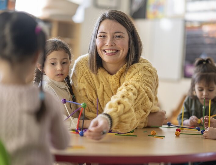 Eine Frau in gelbem Pullover lächelt, während sie mit Kindern spielt, die bunte Bausteine halten und Strukturen bauen.