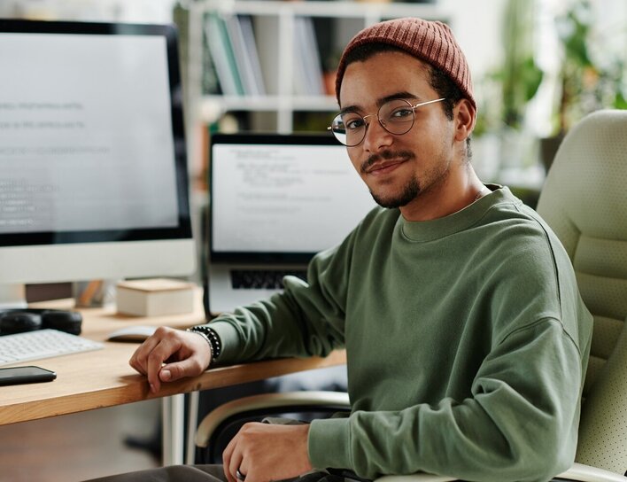 Mann mit Brille und Mütze an Schreibtisch Mann mit Brille und Mütze sitzt an einem Schreibtisch in einem modernen Büro, schaut lächelnd zur Kamera. Im Hintergrund befinden sich zwei Monitore, Bücherregale und Büromaterialien.