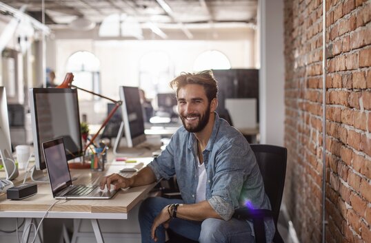 Lächelnder Mann arbeitet am Schreibtisch in modernem Büro mit Backsteinwand