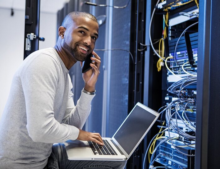 Ein Mann mit Laptop spricht lächelnd am Telefon, während er in einem Serverraum an Netzwerktechnik arbeitet.
