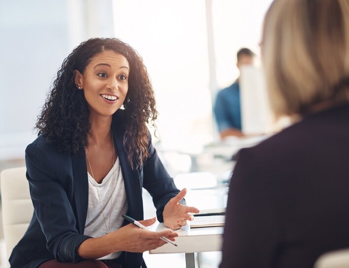 Zwei Frauen sitzen gemeinsam an einem Bürotisch und unterhalten sich.