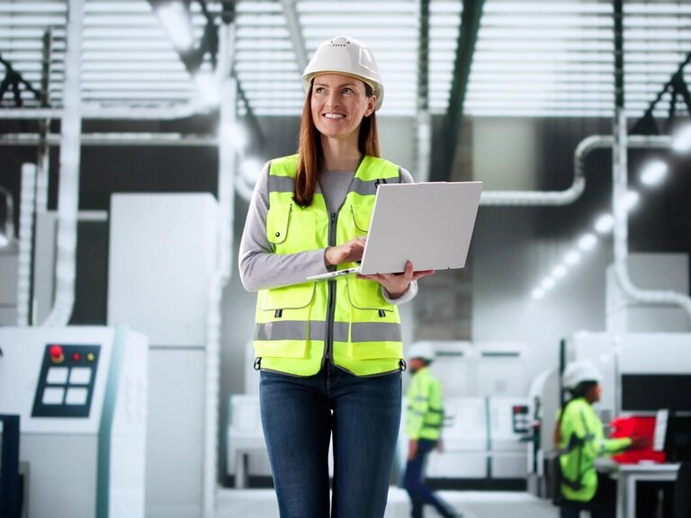 Eine Frau mit Schutzhelm und Warnweste arbeitet mit einem Laptop in einer modernen Produktionshalle.