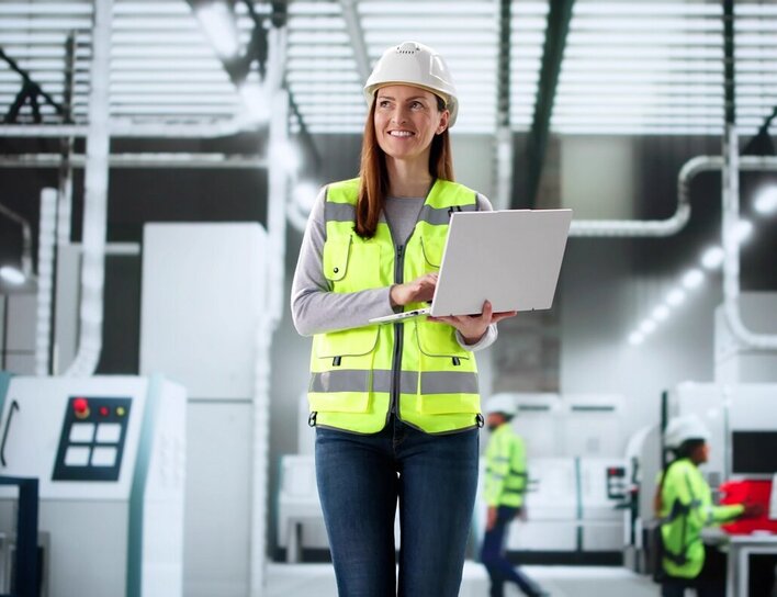Eine Frau mit Schutzhelm und Warnweste arbeitet mit einem Laptop in einer modernen Produktionshalle.