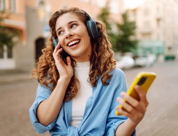 FAQ Studium Frau mit Kopfhörern telefoniert lachend auf der Straße mit ihrem Smartphone.