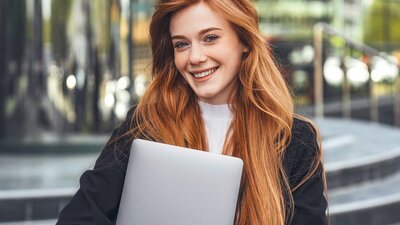 Eine junge Frau mit langen roten Haaren steht vor einem modernen Gebäude und lächelt in die Kamera. Sie trägt ein Business-Outfit und hält ein Laptop im Arm.