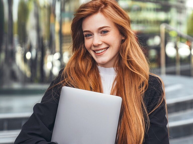 Eine junge Frau mit langen roten Haaren steht vor einem modernen Gebäude und lächelt in die Kamera. Sie trägt ein Business-Outfit und hält ein Laptop im Arm.