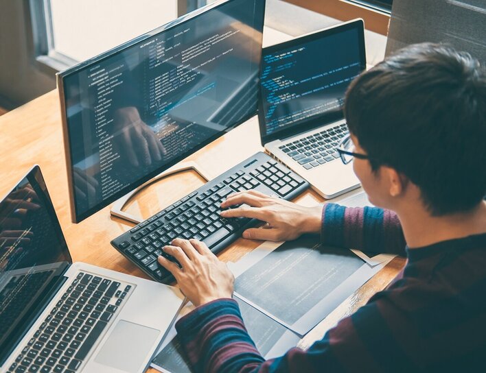 Ein Mann sitzt vor drei Bildschirmen und programmiert etwas.